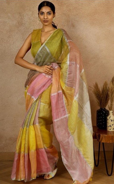 Woman wearing a sleeveless olive-green blouse paired with a soft, pastel-colored linen saree featuring large checks in shades of pink, yellow, white, and green, styled in a traditional drape, standing indoors against a textured beige wall.
